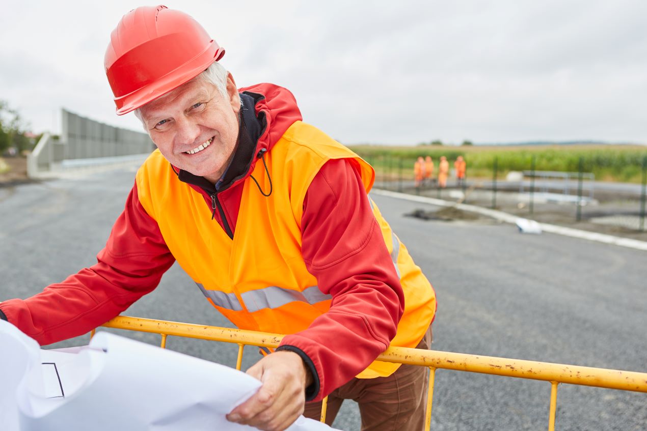 Ein Bauleiter mit rotem Helm und Warnweste überprüft die Planung für neue Fahrbahnmarkierungen auf einer Straßenbaustelle, während Arbeiter im Hintergrund arbeiten.